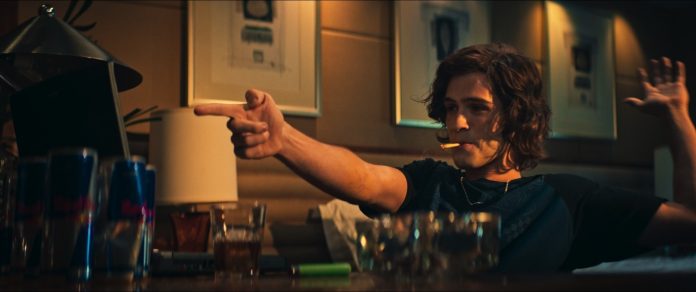 Man with shoulder-length hair sits at a dimly lit bar, cigarette in mouth, pointing off-screen with his right hand and raising his left hand in the air.