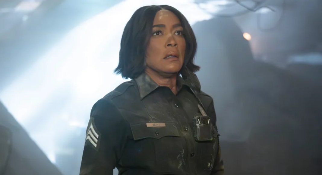 Female officer in a dark uniform with sergeant stripes stands in a smoky, dramatic scene, looking upward with a concerned expression.