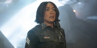 Female officer in a dark uniform with sergeant stripes stands in a smoky, dramatic scene, looking upward with a concerned expression.
