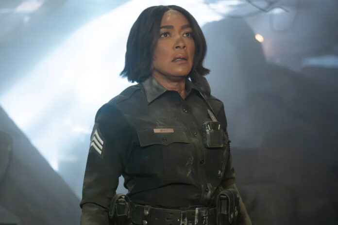 Female officer in a dark uniform with sergeant stripes stands in a smoky, dramatic scene, looking upward with a concerned expression.