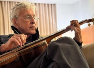 Older man with gray hair and glasses sitting on a couch, playing an acoustic guitar at home.