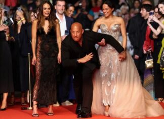 Bald man in a black suit performs a dynamic pose on a red carpet between two women in elegant gowns as fans take photos in the background.