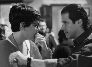 Two people face each other closely in a black-and-white photo; the woman with glasses touches her face while the man extends his arm toward her.