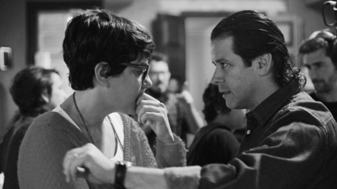 Two people face each other closely in a black-and-white photo; the woman with glasses touches her face while the man extends his arm toward her.