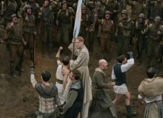 ‘Coward’: Filme de Lukas Dhont é escolhido pela Bélgica para disputar o Oscar 2027 A pale-robed man at the center holds a blue flag as soldiers close in with weapons around him in a chaotic battle on a muddy ground.