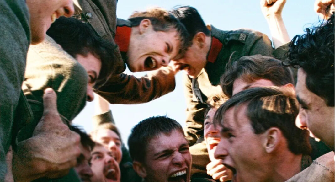 Group of soldiers in green uniforms cheer in a tight circle, fists raised, faces yelling in a moment of intense camaraderie.