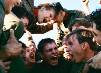 Group of soldiers in green uniforms cheer in a tight circle, fists raised, faces yelling in a moment of intense camaraderie.