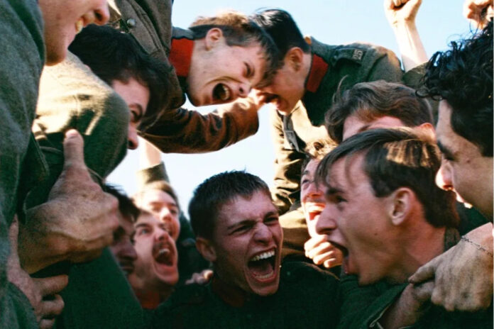 Group of soldiers in green uniforms cheer in a tight circle, fists raised, faces yelling in a moment of intense camaraderie.