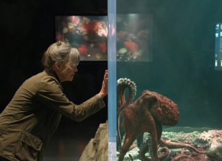 Woman in a brown jacket presses her hands to the glass of an aquarium, watching a large octopus among coral on the other side.