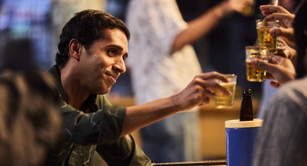 Man toasting with friends at a casual gathering, reaching with a plastic cup of beer toward others; a blue cooler is nearby.