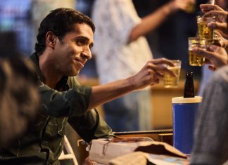 Man toasting with friends at a casual gathering, reaching with a plastic cup of beer toward others; a blue cooler is nearby.