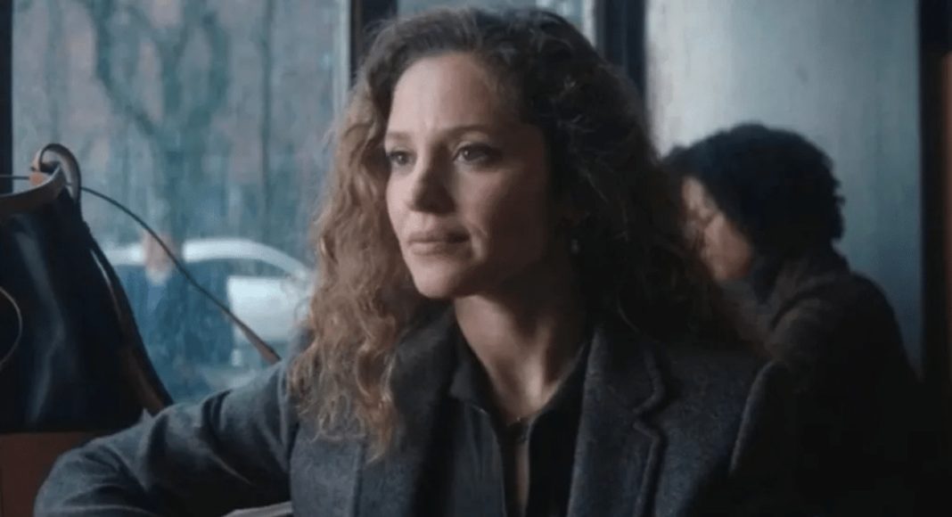 Woman with curly hair in a gray blazer sits at a cafe table, rain visible outside the window.