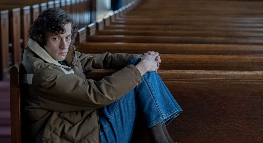 Dominic Sessa os rejeitados (1) Young man sitting alone in a church pew, arms wrapped around his knees, looking at the camera with a somber expression.