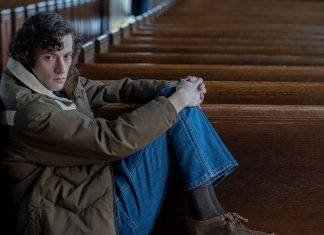 Young man sitting alone in a church pew, arms wrapped around his knees, looking at the camera with a somber expression.