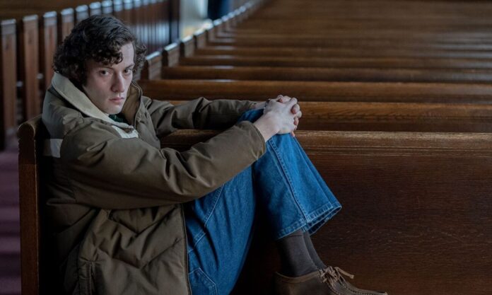 Young man sitting alone in a church pew, arms wrapped around his knees, looking at the camera with a somber expression.