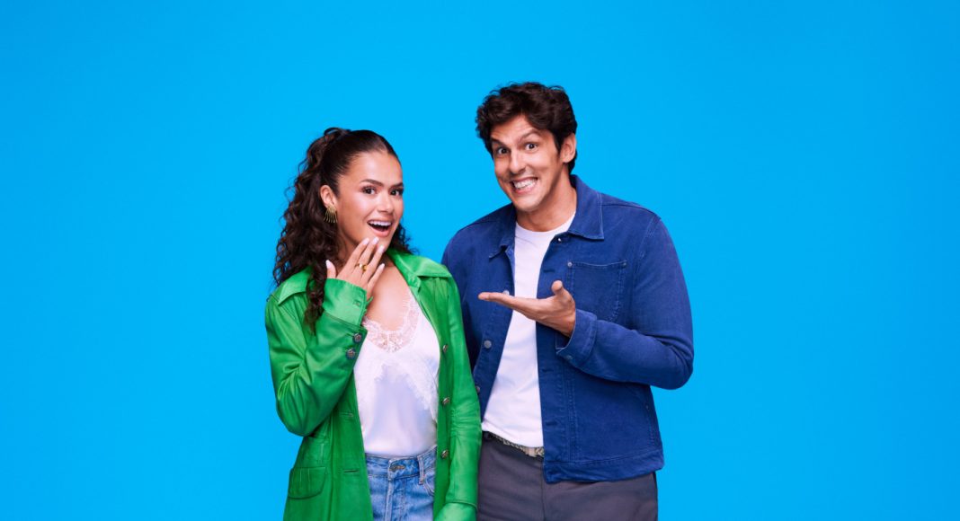 Two friends pose happily against a bright blue background, the woman in a green coat and the man in a denim jacket smiling at the camera with an inviting gesture.
