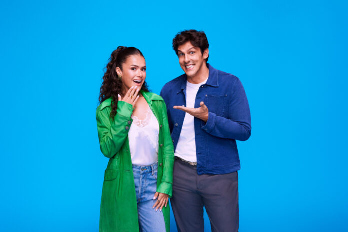 Two friends pose happily against a bright blue background, the woman in a green coat and the man in a denim jacket smiling at the camera with an inviting gesture.