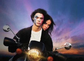 Young couple riding a motorcycle at sunset; the man has blue face paint and the woman leans close behind him, both looking ahead.