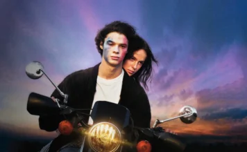Young couple riding a motorcycle at sunset; the man has blue face paint and the woman leans close behind him, both looking ahead.