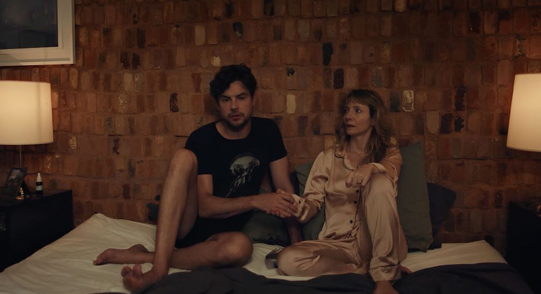 Couple sits on a bed in a dim brick-walled room, holding hands and looking toward each other.