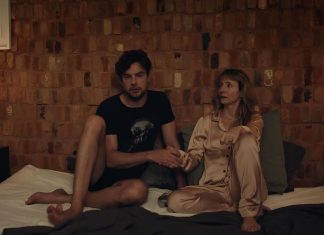 ‘Eclipse’: Suspense INTENSO para te deixar reflexivo está em cartaz nos cinemas Couple sits on a bed in a dim brick-walled room, holding hands and looking toward each other.