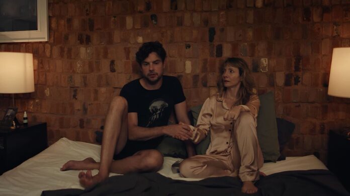 Couple sits on a bed in a dim brick-walled room, holding hands and looking toward each other.