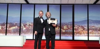 Two men in tuxedos pose on a red-carpet stage; one holds an award plaque against a cityscape backdrop.