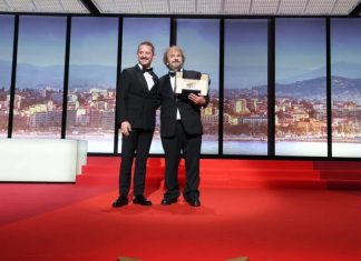 Two men in tuxedos pose on a red-carpet stage; one holds an award plaque against a cityscape backdrop.