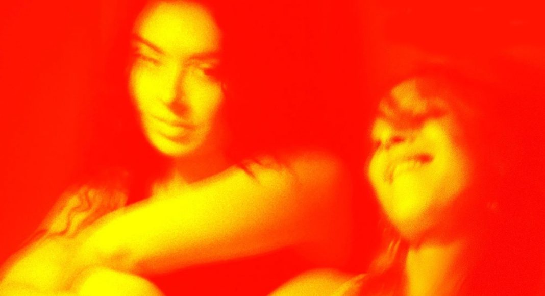 Two people posing and smiling together in a warm red-toned photo with a tattoo on one arm visible on the left