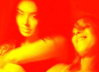 Two people posing and smiling together in a warm red-toned photo with a tattoo on one arm visible on the left