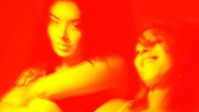 Two people posing and smiling together in a warm red-toned photo with a tattoo on one arm visible on the left