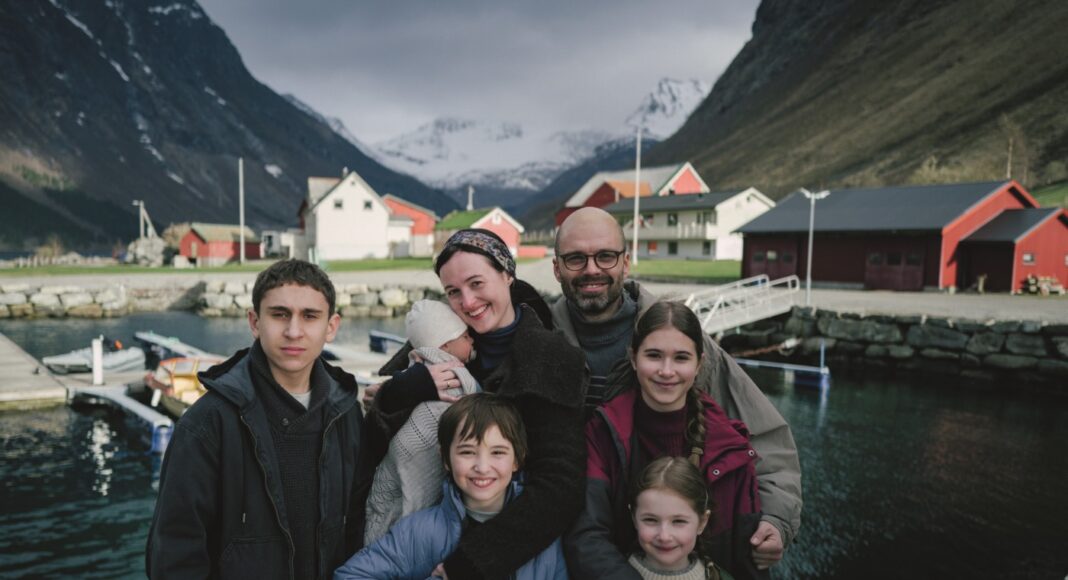 A seven-member family posing together by a small harbor in a Norwegian village, with colorful red and white houses and snow-covered mountains in the background.