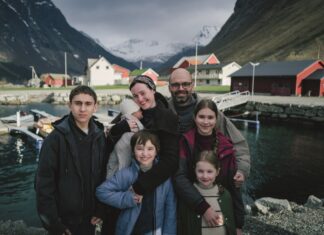 A seven-member family posing together by a small harbor in a Norwegian village, with colorful red and white houses and snow-covered mountains in the background.
