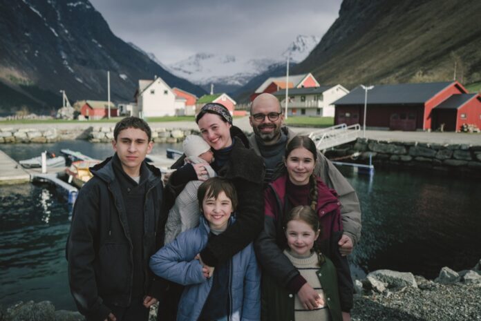 A seven-member family posing together by a small harbor in a Norwegian village, with colorful red and white houses and snow-covered mountains in the background.