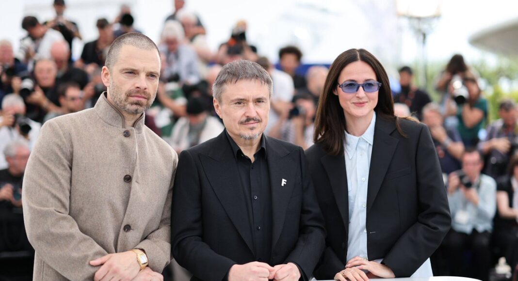Three formally dressed individuals pose behind a white table at a press event, with photographers in the background.