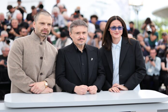 Three formally dressed individuals pose behind a white table at a press event, with photographers in the background.