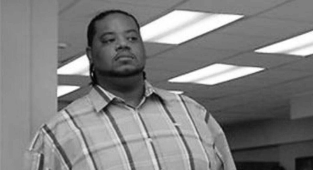 Black-and-white photo of a man in a plaid shirt standing in an office, looking straight ahead with a serious expression.