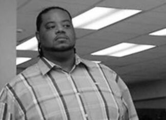Grizzwald “Grizz” Chapman, ator de ’30 Rock’, morre aos 52 anos Black-and-white photo of a man in a plaid shirt standing in an office, looking straight ahead with a serious expression.