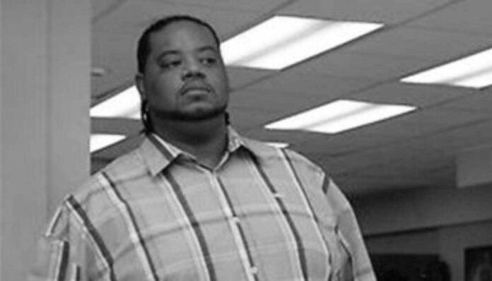 Black-and-white photo of a man in a plaid shirt standing in an office, looking straight ahead with a serious expression.