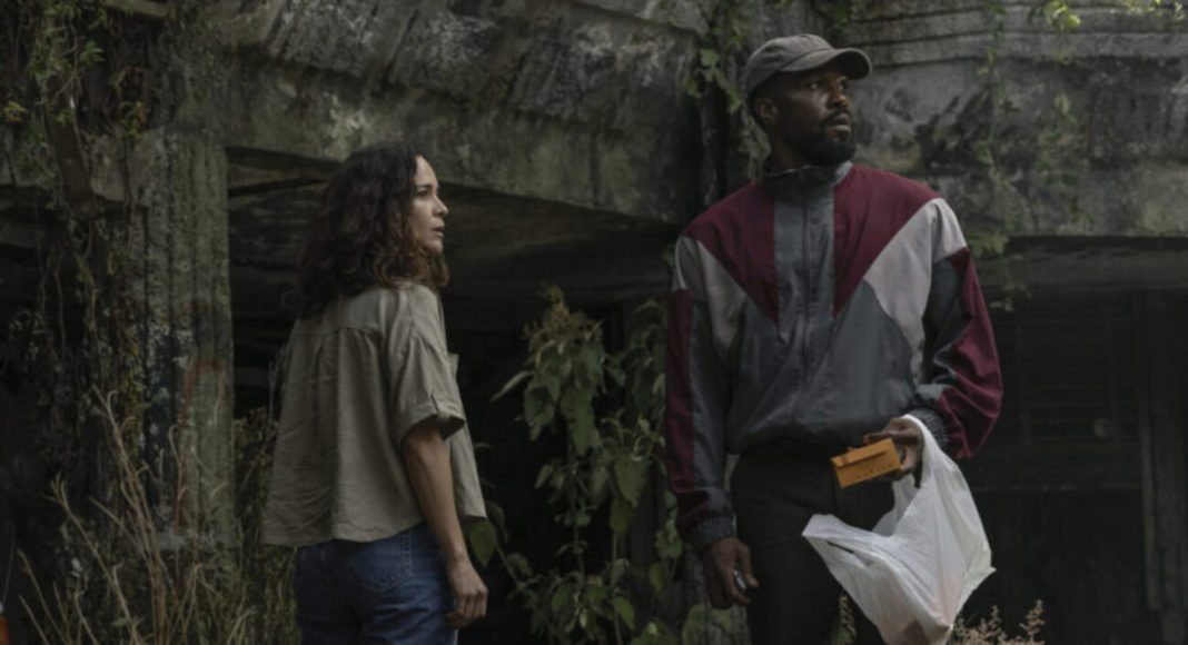 Two adults stand outside by a mossy concrete wall; the man in a color-block windbreaker holds a yellow envelope and a plastic bag while the woman watches nearby.