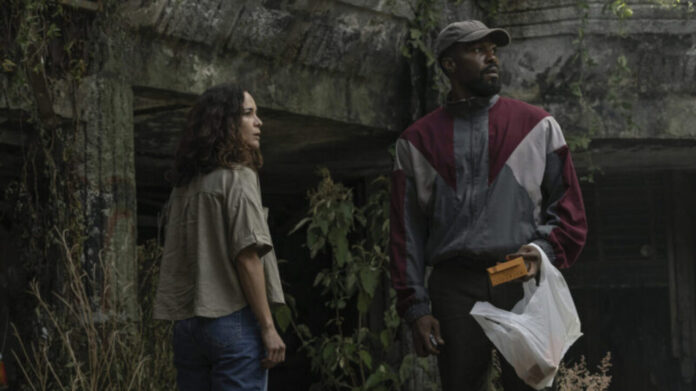 Two adults stand outside by a mossy concrete wall; the man in a color-block windbreaker holds a yellow envelope and a plastic bag while the woman watches nearby.