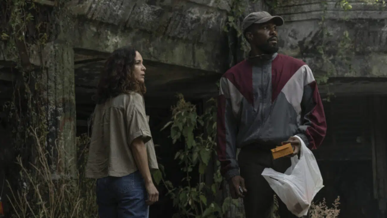Two adults stand outside by a mossy concrete wall; the man in a color-block windbreaker holds a yellow envelope and a plastic bag while the woman watches nearby.