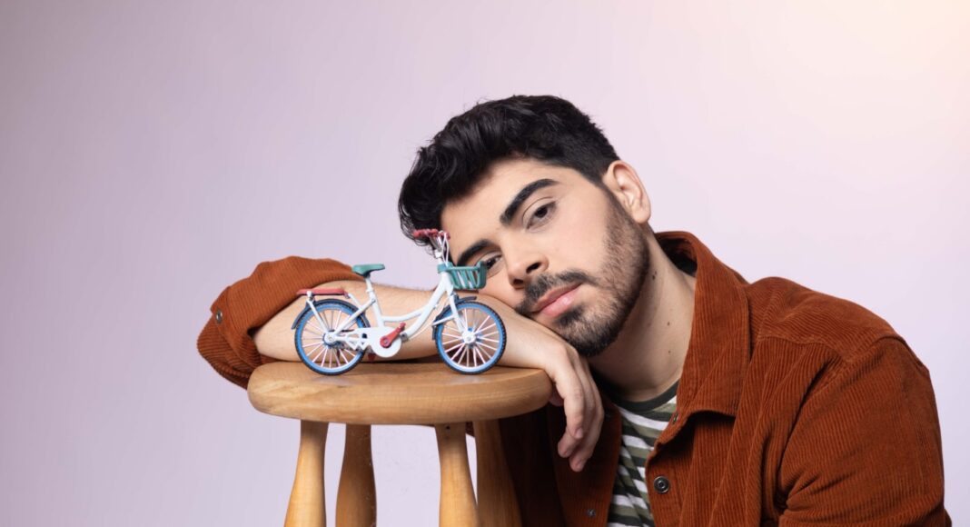 IMG_9280 Young man with dark hair and beard rests his head on a wooden stool, a small blue toy bicycle sits beside his arm on the stool.