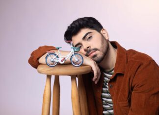 Young man with dark hair and beard rests his head on a wooden stool, a small blue toy bicycle sits beside his arm on the stool.