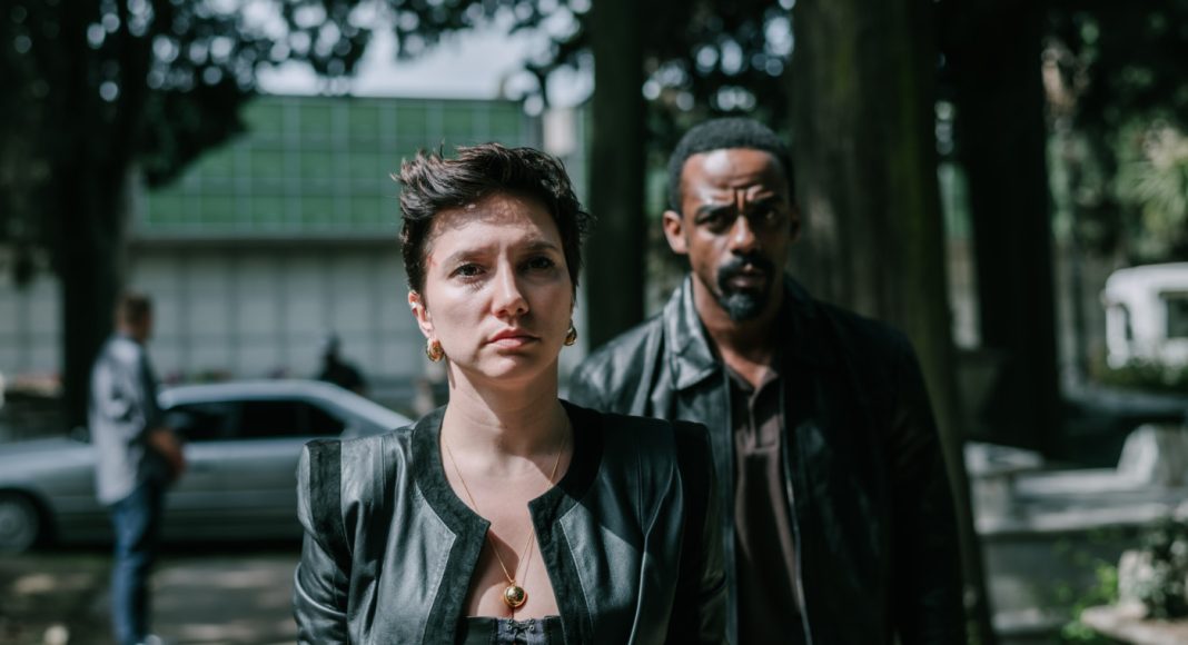 Woman in a black leather jacket at the forefront with a serious expression; a man in a dark jacket stands behind her on a tree-lined urban street with a blurred car in the background.