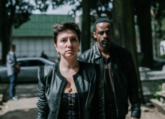 Woman in a black leather jacket at the forefront with a serious expression; a man in a dark jacket stands behind her on a tree-lined urban street with a blurred car in the background.