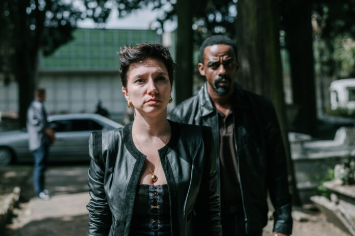 Woman in a black leather jacket at the forefront with a serious expression; a man in a dark jacket stands behind her on a tree-lined urban street with a blurred car in the background.