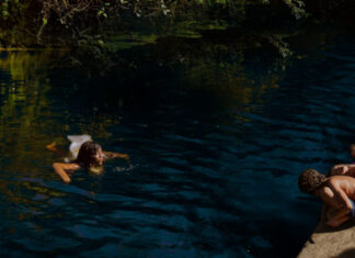 Person swimming in a dark blue lake near the rocky shore while others relax on a colorful towel at the bank.
