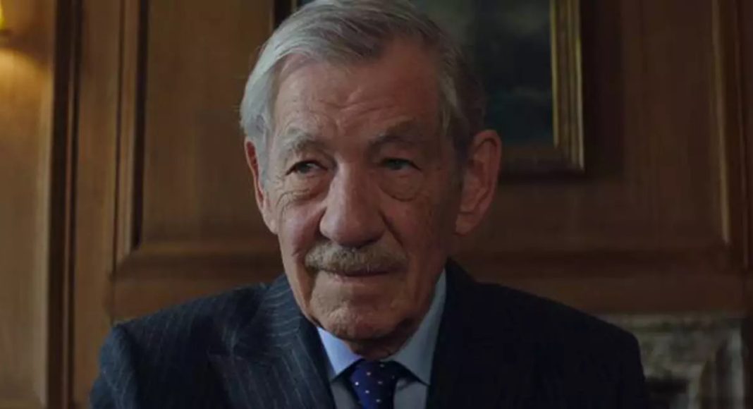 An older man with gray hair and a mustache wearing a dark suit and tie, seated in a wood-paneled room.