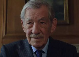 An older man with gray hair and a mustache wearing a dark suit and tie, seated in a wood-paneled room.
